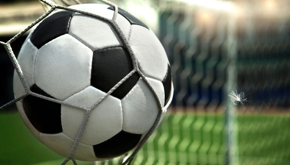 Bola de futebol presa na rede do gol durante partida em campo verde, destacando emoção do esporte e momento decisivo.