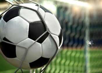 Bola de futebol presa na rede do gol durante partida em campo verde, destacando emoção do esporte e momento decisivo.