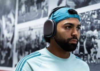 Gabigol alfineta Roger Flores: 'Olha quem fala agora!' 31 Homem de boné azul e fones de ouvido grandes caminha em frente a uma parede com fotos antigas em preto e branco.