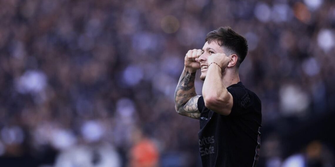 Jogador de futebol em campo celebra com gesto animado, cercado por torcida em estádio lotado, destacando emoção do esporte.