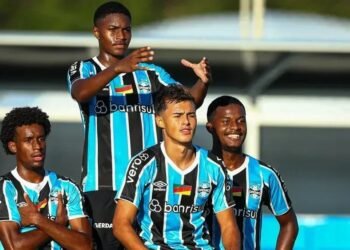 Jogadores do Grêmio com uniformes listrados comemoram em campo sob céu claro, expressando foco e entusiasmo.