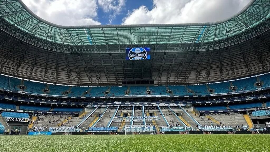 Estádio de futebol com arquibancadas azuis e grandes telões, gramado verde em primeiro plano sob céu parcialmente nublado.