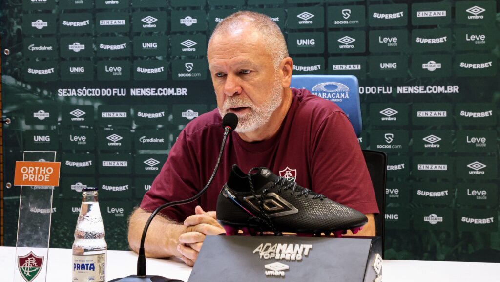 Homem em coletiva, ao microfone, com chuteira preta sobre mesa. Fundo verde com logotipos e garrafa de água à frente.