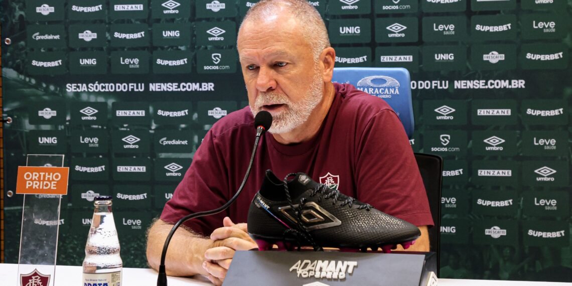 Homem em coletiva, ao microfone, com chuteira preta sobre mesa. Fundo verde com logotipos e garrafa de água à frente.
