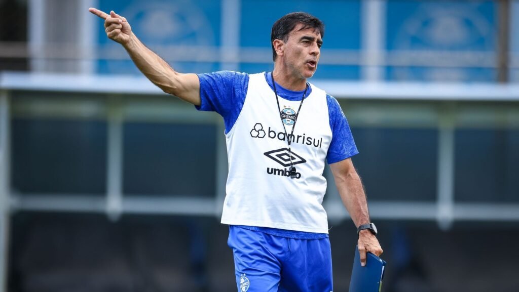 Treinador do Grêmio dá instruções energéticas em campo de treino, vestindo uniforme do time gaúcho com expressão focada.
