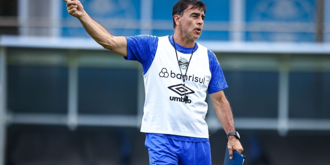 Treinador do Grêmio dá instruções energéticas em campo de treino, vestindo uniforme do time gaúcho com expressão focada.
