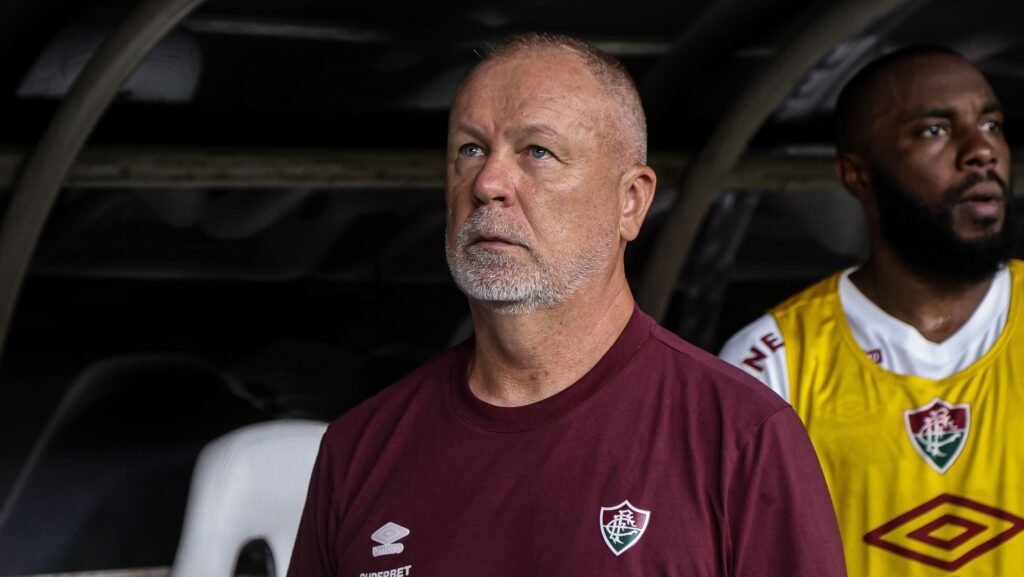 Treinador de futebol do Fluminense observa atentamente o campo durante a partida, ao lado de jogador no banco de reservas.