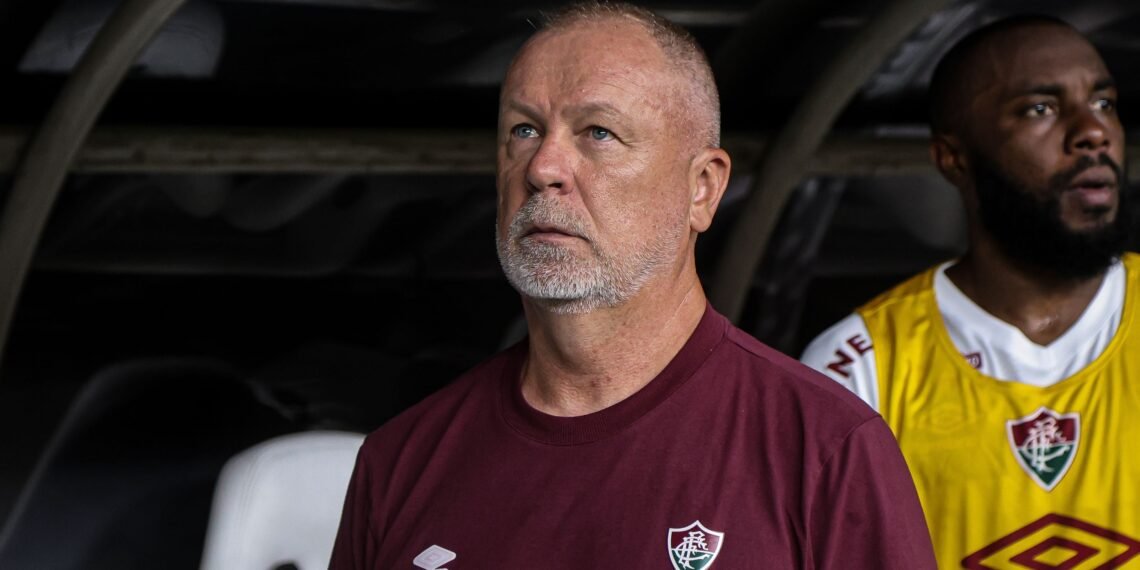 Treinador de futebol do Fluminense observa atentamente o campo durante a partida, ao lado de jogador no banco de reservas.