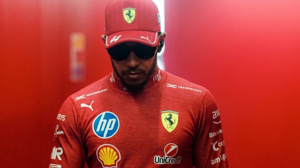 Piloto de Fórmula 1 veste uniforme vermelho da Ferrari com logotipos de patrocinadores, usando boné e óculos escuros.