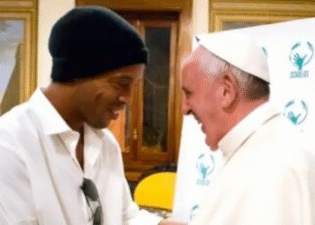 Dois homens sorridentes apertam as mãos em um ambiente formal, em frente a um painel com o logo Scholas.