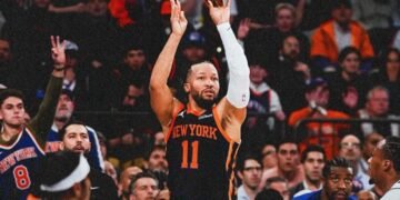 Jogador do New York Knicks arremessa bola de basquete em quadra lotada, enquanto torcida observa atenta a jogada decisiva.