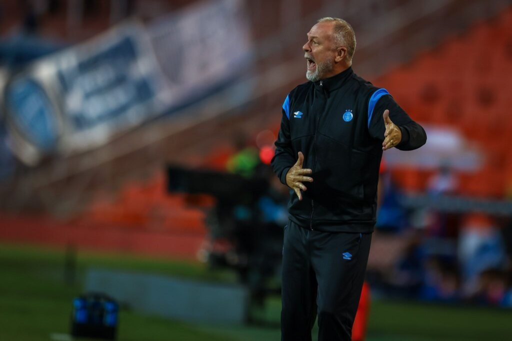 Técnico de futebol gesticula intensamente à beira do campo, durante jogo em estádio, demonstrando tática para equipe.