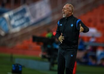 Técnico de futebol gesticula intensamente à beira do campo, durante jogo em estádio, demonstrando tática para equipe.