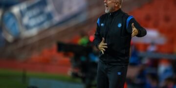 Técnico de futebol gesticula intensamente à beira do campo, durante jogo em estádio, demonstrando tática para equipe.