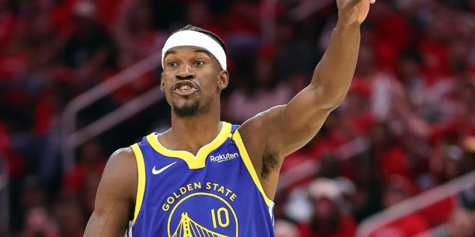 Jogador de basquete do Golden State Warriors usando faixa na cabeça gesticula em quadra durante jogo, torcida ao fundo.