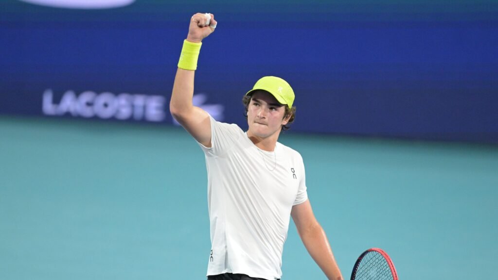 Tenista celebra com punho erguido, usando boné e munhequeira amarela, em quadra de piso azul.