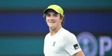 Tenista sorridente usa boné verde e camiseta branca durante partida em quadra, destacando-se em evento esportivo.