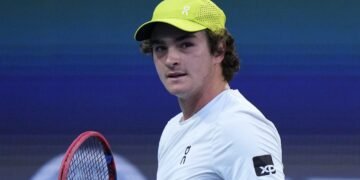 Jovem tenista com boné amarelo e raquete de tênis, vestido com camiseta branca, durante partida em quadra azul.