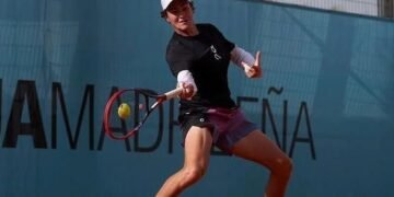 Jogador de tênis se prepara para rebater uma bola em quadra, usando boné e roupa esportiva, com uma parede azul ao fundo.