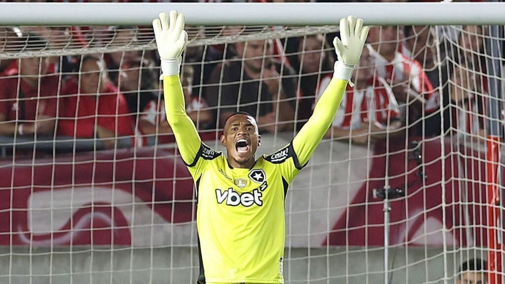 Goleiro comemora defesa em jogo de futebol, vestindo uniforme amarelo vibrante, em frente a torcida empolgada no estádio.