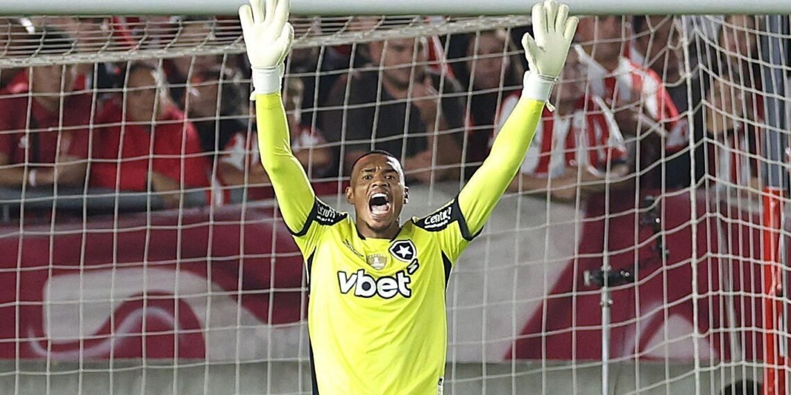 Goleiro comemora defesa em jogo de futebol, vestindo uniforme amarelo vibrante, em frente a torcida empolgada no estádio.