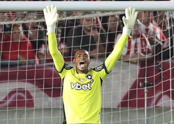 Goleiro comemora defesa em jogo de futebol, vestindo uniforme amarelo vibrante, em frente a torcida empolgada no estádio.