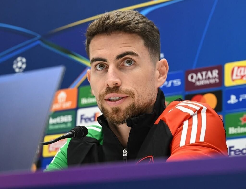 Homem com barba em coletiva de imprensa de futebol, com microfone e fundo com logotipos de patrocinadores de campeonato.