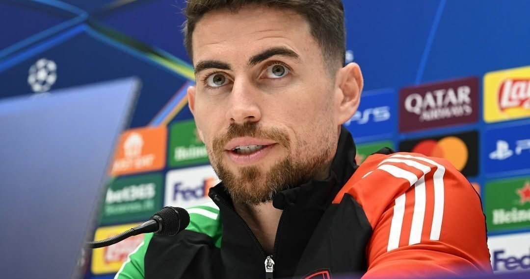 Homem com barba em coletiva de imprensa de futebol, com microfone e fundo com logotipos de patrocinadores de campeonato.