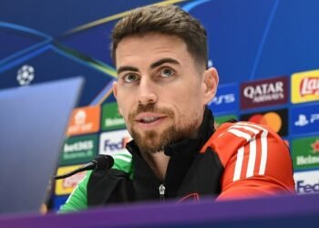 Homem com barba em coletiva de imprensa de futebol, com microfone e fundo com logotipos de patrocinadores de campeonato.