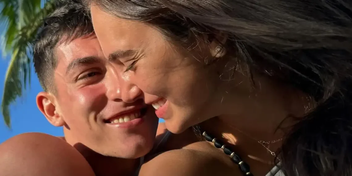 Casal sorridente em clima de romance sob céu azul, com palmeira ao fundo, destacando sensação de felicidade e amor.
