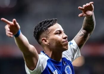 Jogador de futebol comemora com os braços abertos, vestindo uniforme azul e branco, em campo durante partida emocionante.