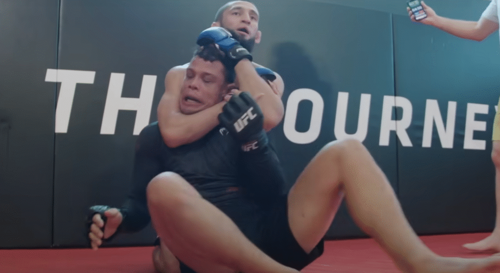 Dois lutadores treinam no octógono; um aplica mata-leão no outro em movimento técnico de finalização no MMA.