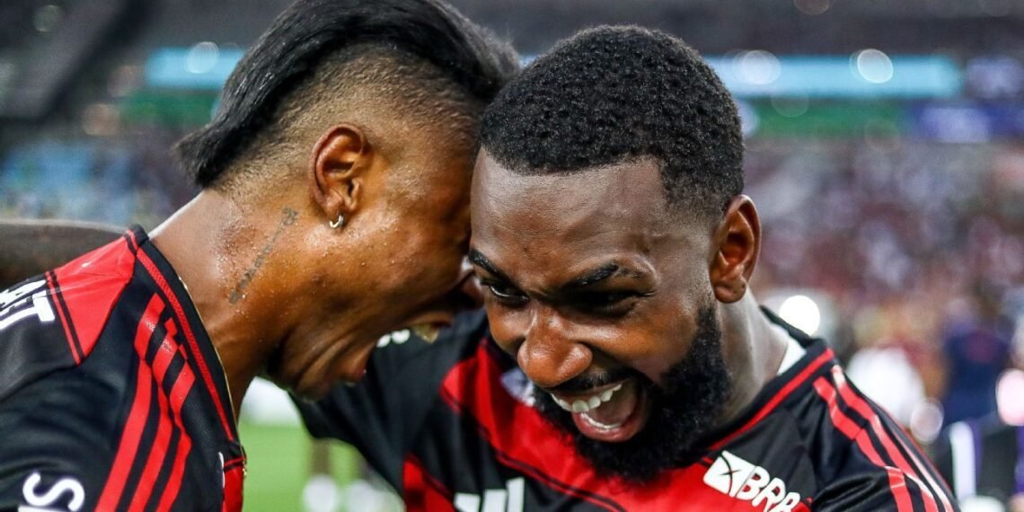 Jogadores de futebol do Flamengo comemoram intensamente, vestindo uniformes vermelhos e pretos, em um estádio vibrante.