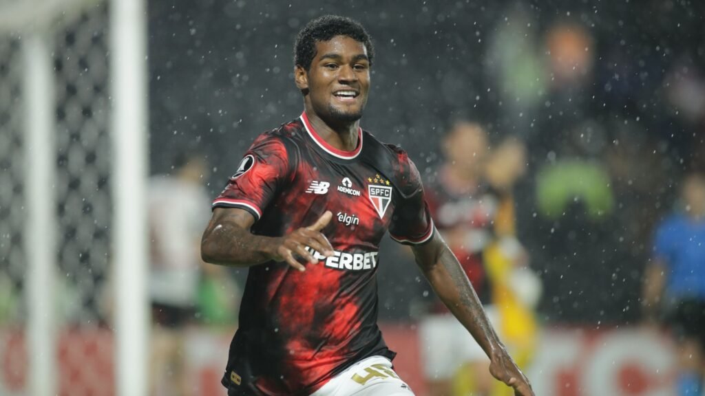 Jogador de futebol do São Paulo celebra gol em campo, sob chuva, vestindo uniforme vermelho e preto com patrocínios visíveis.