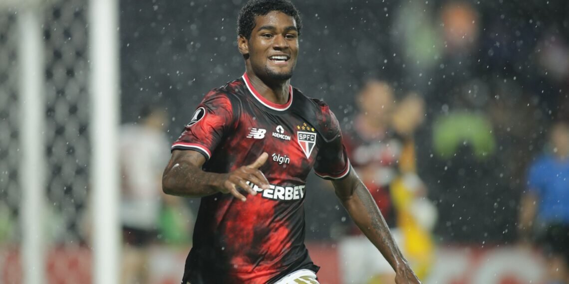 Jogador de futebol do São Paulo celebra gol em campo, sob chuva, vestindo uniforme vermelho e preto com patrocínios visíveis.
