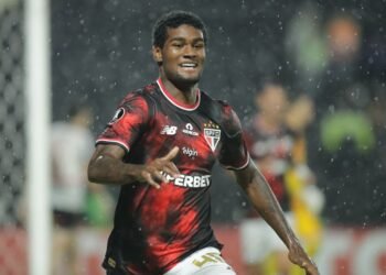 Lucas Ferreira: Protagonista do Gol do São Paulo contra o Libertad 34 Jogador de futebol do São Paulo celebra gol em campo, sob chuva, vestindo uniforme vermelho e preto com patrocínios visíveis.