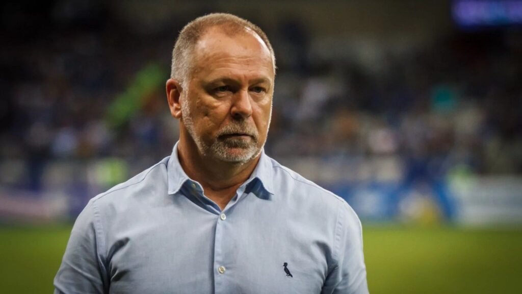 Homem de camisa azul claro observa atentamente um campo de futebol desfocado ao fundo, transmitindo foco e concentração.