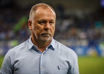 Homem de camisa azul claro observa atentamente um campo de futebol desfocado ao fundo, transmitindo foco e concentração.