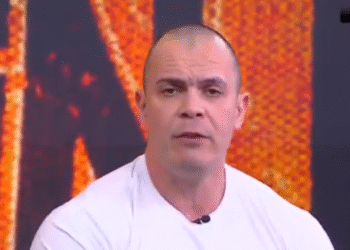 Um homem de camiseta branca participa de um programa de TV, com hashtag do programa no canto superior direito.