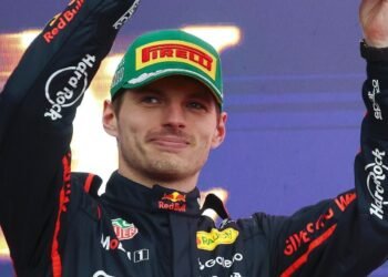 Max Verstappen troca Red Bull por equipe rival em 2024! 31 Piloto de corrida celebra vitória em pódio, usando macacão de equipe e boné patrocinado, sorrindo e levantando os braços.