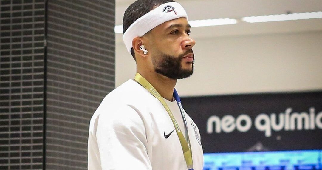 Homem usando faixa de cabeça e fones de ouvido caminha em ambiente fechado, vestindo uniforme esportivo com logotipo visível.