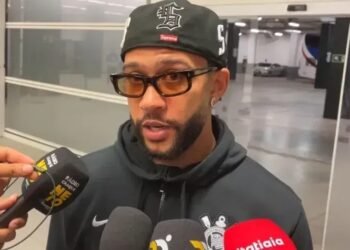 Homem com boné preto e óculos de sol fala para a imprensa com vários microfones; cenário de estacionamento ao fundo.