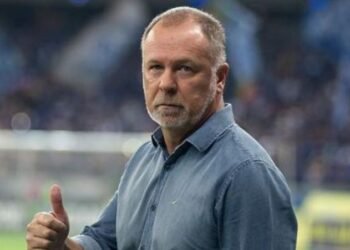 Treinador em campo de futebol, vestindo camisa azul, faz sinal de positivo, com expressão séria e torcedores ao fundo.