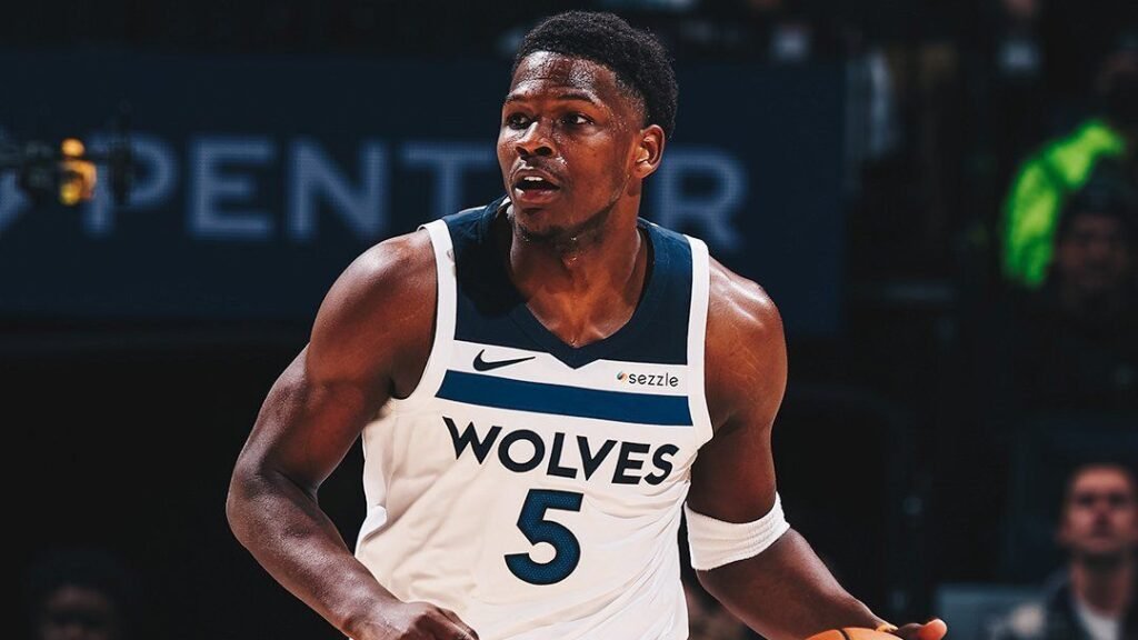 Jogador de basquete em ação com camisa Wolves, durante jogo, em quadra com iluminação intensa e público ao fundo.