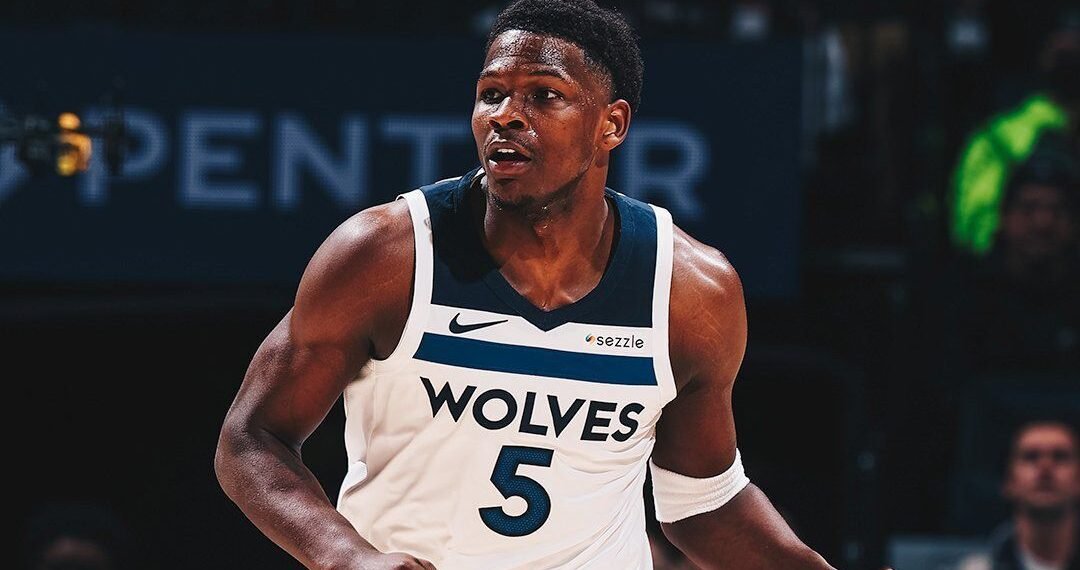 Jogador de basquete em ação com camisa Wolves, durante jogo, em quadra com iluminação intensa e público ao fundo.