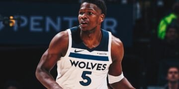 Jogador de basquete em ação com camisa Wolves, durante jogo, em quadra com iluminação intensa e público ao fundo.