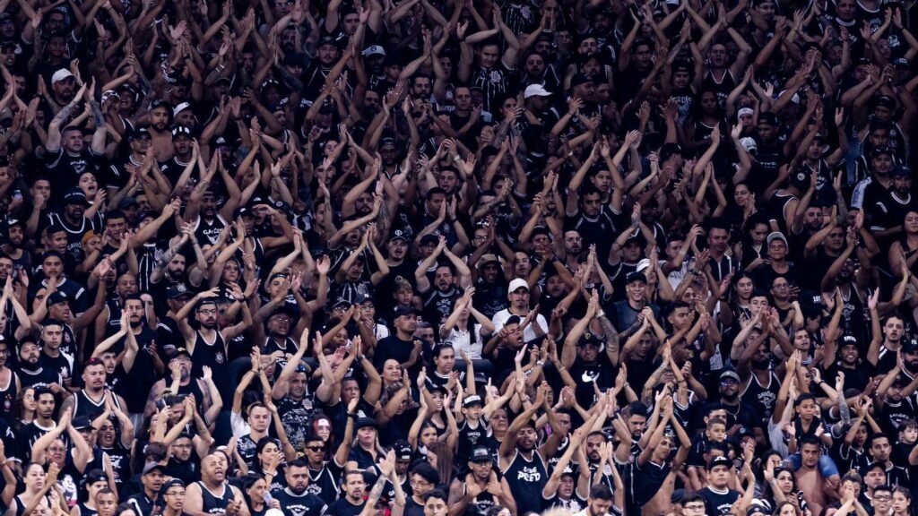 Torcida vibrante em estádio de futebol, uniformizada em preto e branco, aplaude e celebra o time durante o jogo.