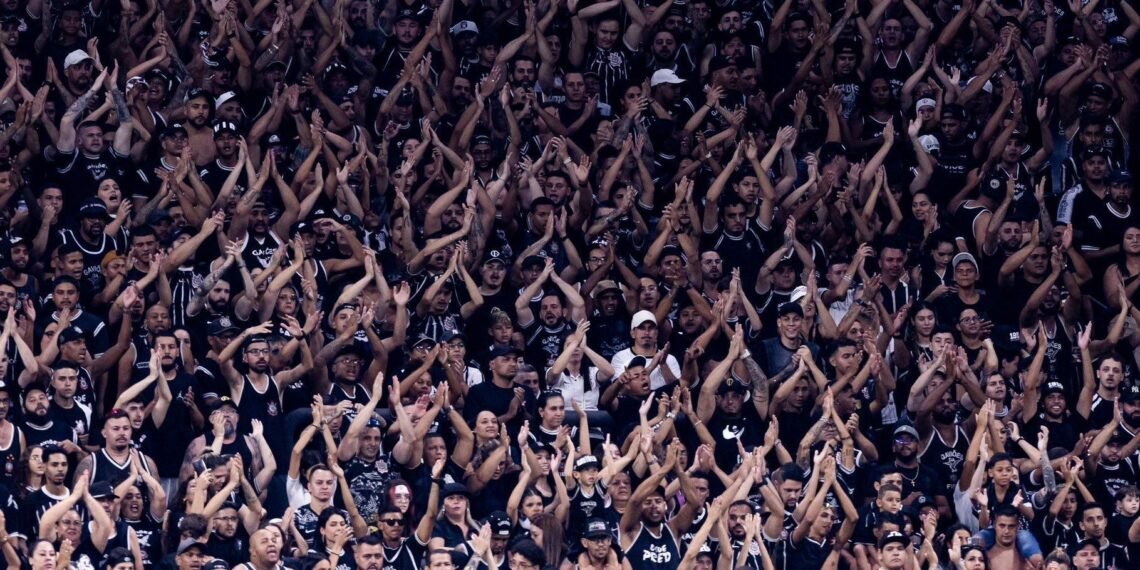 Torcida vibrante em estádio de futebol, uniformizada em preto e branco, aplaude e celebra o time durante o jogo.