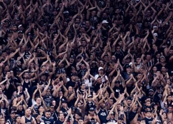 Torcida vibrante em estádio de futebol, uniformizada em preto e branco, aplaude e celebra o time durante o jogo.