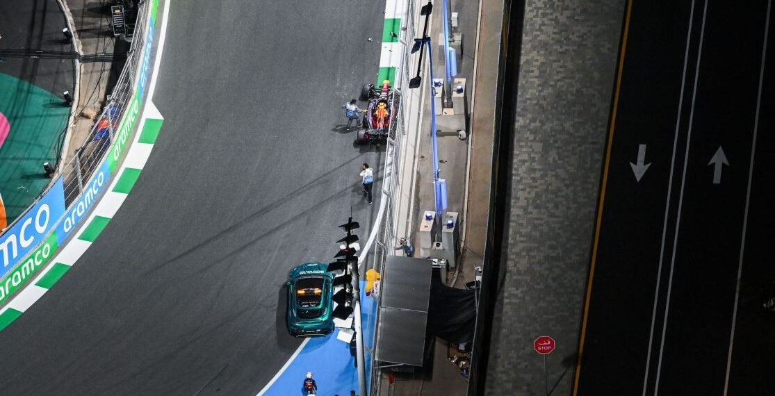 Vista aérea de uma pista de corrida com um carro parado, equipes próximas ao veículo e sinalização de trânsito visível.